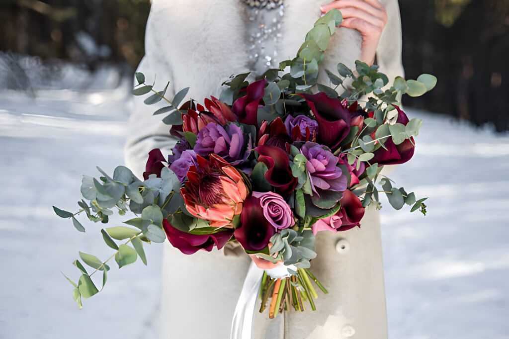 Close-up of a bride holding a winter wedding bouquet with deep jewel-toned flowers like burgundy calla lilies, purple roses, and a protea, accented with eucalyptus greenery, against a soft, snowy background.